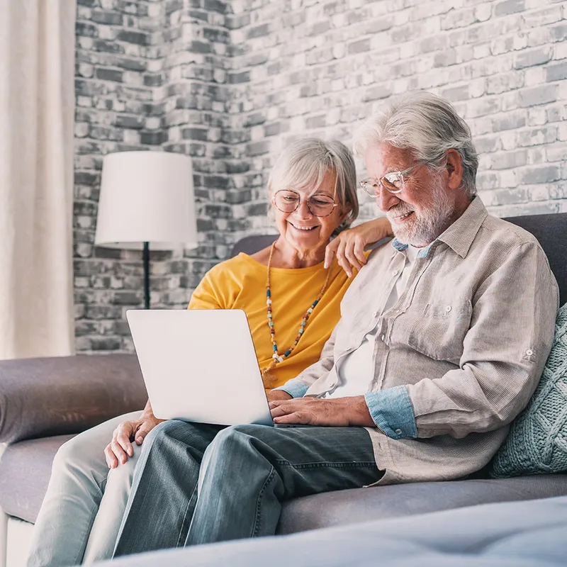 Zwei lächelnde Personen im Wohnzimmer auf Sofa mit Laptop